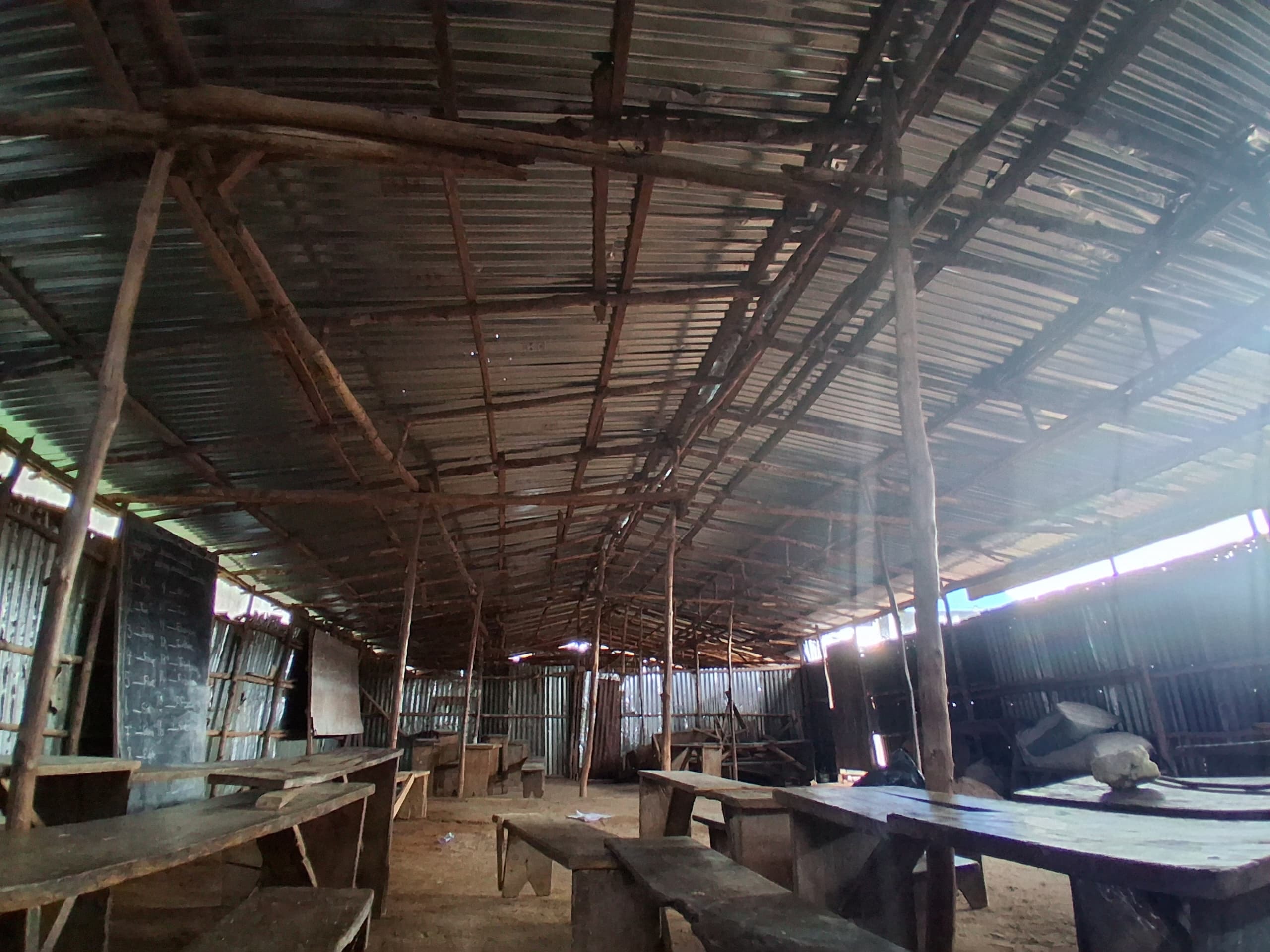 Wide interior view of the classroom showing wooden beam roof structure, zinc walls, and long wooden benches
