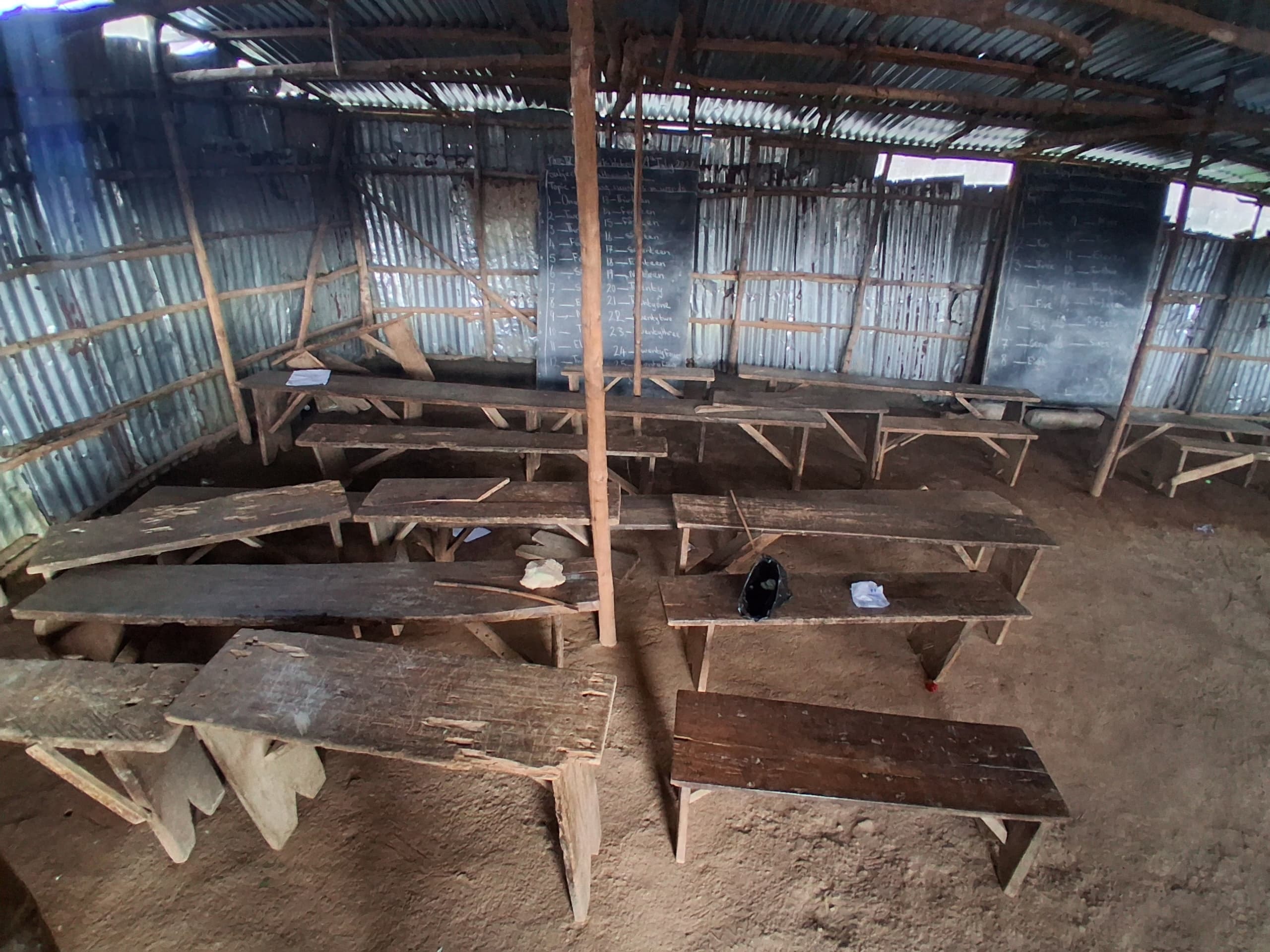 Close view of wooden classroom benches and desks arranged on a dirt floor beneath a zinc roof